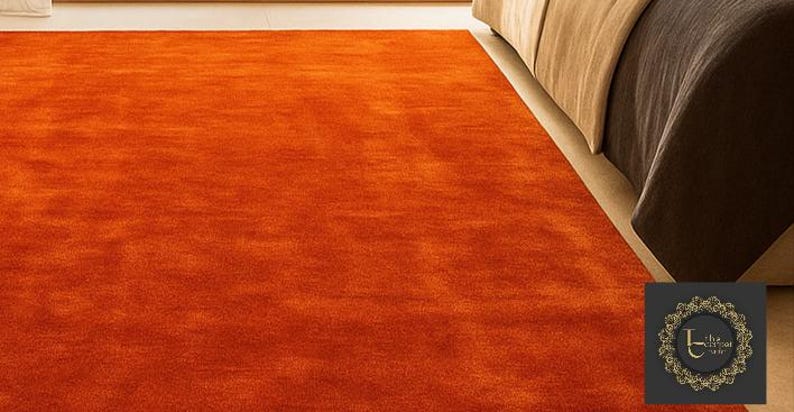 Op de afbeelding: Een feloranje vloerkleed met een zacht, getextureerd oppervlak. Het vloerkleed staat naast een bed met een bruin dekbed en een beige bedrok. De kleur van het vloerkleed voegt warmte toe aan de kamer.