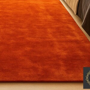 Op de afbeelding: Een levendig oranje tapijt met een zacht, getextureerd oppervlak. Het tapijt ligt op een lichtgekleurde vloer, met een deel van een bed op de achtergrond. De kleur van het tapijt is uniform, wat een warme uitstraling cre&euml;ert.