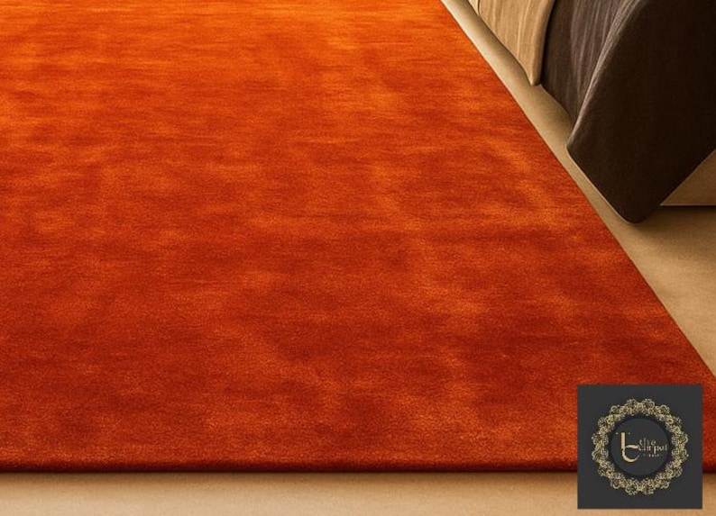 Op de afbeelding: Een levendig oranje tapijt met een zacht, getextureerd oppervlak. Het tapijt ligt op een lichtgekleurde vloer, met een deel van een bed op de achtergrond. De kleur van het tapijt is uniform, wat een warme uitstraling cre&euml;ert.