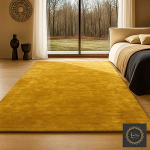 May include: A large, rectangular mustard-yellow rug in a bedroom setting. The rug is placed on a wooden floor, with a bed and large window in the background. Decorative vases and a wall hanging are also visible.