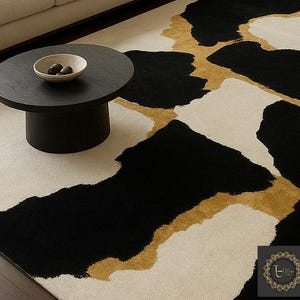 May include: A modern area rug with an abstract design. The rug features large black and off-white shapes with gold accents. A dark round coffee table with a bowl of decorative objects sits on the rug.