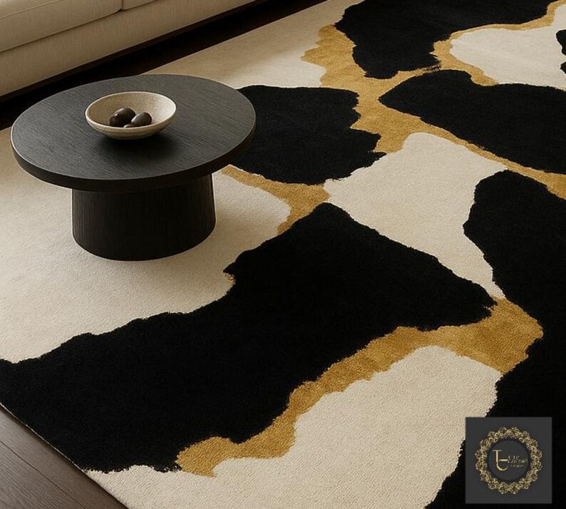 May include: A modern area rug with an abstract design. The rug features large black and off-white shapes with gold accents. A dark round coffee table with a bowl of decorative objects sits on the rug.