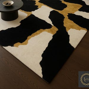 May include: A modern area rug with an abstract design. The rug features large, irregular shapes in black, cream, and gold. The rug is placed on a dark wood floor, with a small black table and a bowl visible in the background.
