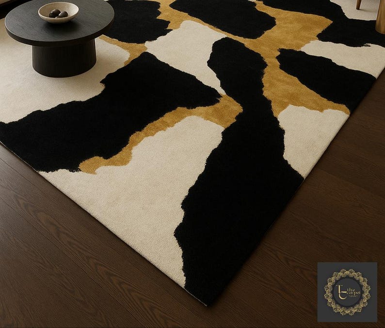 May include: A modern area rug with an abstract design. The rug features large, irregular shapes in black, cream, and gold. The rug is placed on a dark wood floor, with a small black table and a bowl visible in the background.