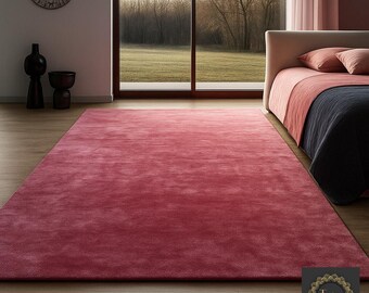 May include: A large, rectangular, solid-colored rug in a deep rose hue dominates the foreground. The rug is placed on a wooden floor, and a bed with rose-colored bedding is visible in the background. The room has a large window.