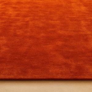 Op de afbeelding: Een groot, rechthoekig oranje tapijt met een zacht, getextureerd oppervlak. Het tapijt is effen van kleur en lijkt gemaakt te zijn van een zacht materiaal. Het tapijt ligt op een lichtgekleurde vloer. Een logo staat rechtsonder.