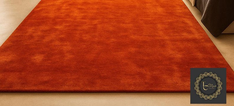 Op de afbeelding: Een groot, rechthoekig oranje tapijt met een zacht, getextureerd oppervlak. Het tapijt is effen van kleur en lijkt gemaakt te zijn van een zacht materiaal. Het tapijt ligt op een lichtgekleurde vloer. Een logo staat rechtsonder.