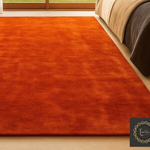 Op de afbeelding: Een groot, rechthoekig, oranje tapijt met een zacht, getextureerd oppervlak. Het tapijt ligt op een lichtgekleurde vloer, met op de achtergrond gedeeltelijk een bed zichtbaar. De kleur van het tapijt voegt warmte toe aan de kamer.