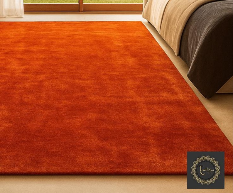 Op de afbeelding: Een groot, rechthoekig, oranje tapijt met een zacht, getextureerd oppervlak. Het tapijt ligt op een lichtgekleurde vloer, met op de achtergrond gedeeltelijk een bed zichtbaar. De kleur van het tapijt voegt warmte toe aan de kamer.