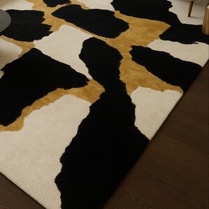 May include: A large area rug with a modern design. The rug features a cream-colored background with large, irregular shapes in black and gold. The rug is placed on a dark wood floor.