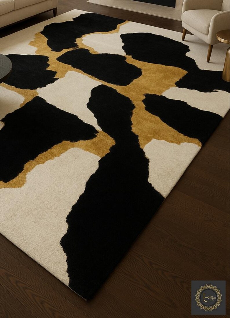 May include: A large area rug with a modern design. The rug features a cream-colored background with large, irregular shapes in black and gold. The rug is placed on a dark wood floor.