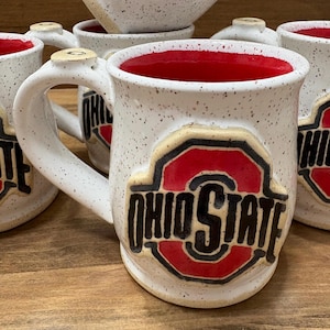 May include: White ceramic mugs with a speckled finish and red interiors. Each mug features the Ohio State logo in red and black. The mugs have a curved handle and are suitable for hot or cold beverages.