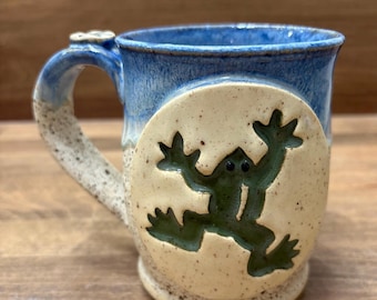 May include: Ceramic mug with a blue rim and handle, featuring a cream-colored body with a green frog design. The handle has a small flower detail. The mug is set on a wooden surface.