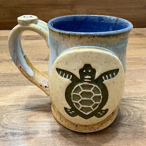 May include: A handmade ceramic mug with a turtle design. The mug features a light blue interior and a tan exterior with a turtle design on the side. The handle has a small, round accent. The mug is approximately 4 inches tall.