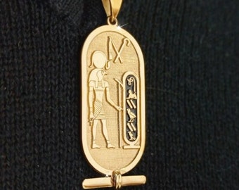 May include: Gold-colored pendant necklace featuring an engraved Egyptian hieroglyphic design. The pendant is oval-shaped with a detailed depiction of a figure and hieroglyphs. The necklace has a delicate chain.