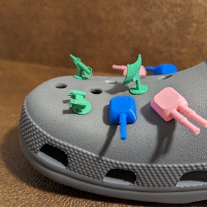 May include: A gray clog with five small, colorful, 3D printed figurines attached to it. The figurines are shaped like cannons and have different colored bases: green, blue, pink, and red.