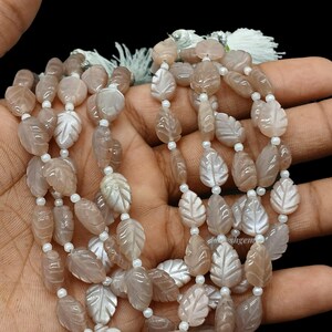 May include: A strand of light peach-colored moonstone beads carved into leaf shapes, interspersed with small white round spacer beads. The gemstone leaves feature a smooth, polished finish with visible veining details and a subtle iridescent sheen.