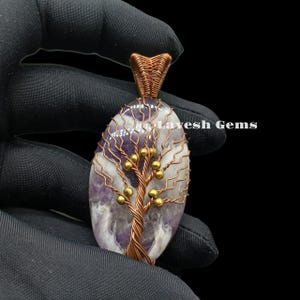 Op de afbeelding: Een hanger van paarse amethyst met een boomontwerp van koperdraad en gouden kralen. De hanger wordt in een hand gehouden.