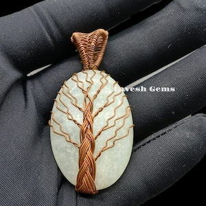 May include: A copper wire tree of life pendant with a white, sparkly, oval-shaped gemstone. The pendant is held in a hand.