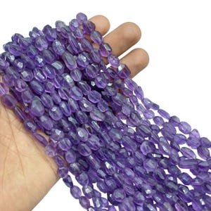 May include: A handful of faceted, oval-shaped amethyst beads. The beads are a deep purple color and appear to be strung together. The beads are held in a person's hand against a white background. These beads are suitable for jewelry making.