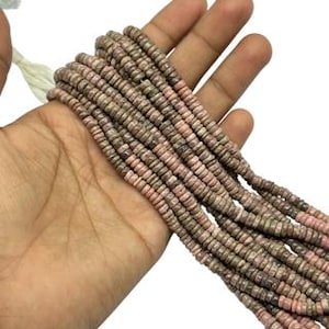 May include: A strand of small, flat, circular beads in alternating shades of dusty pink and muted brown. The beads are strung on a thin white cord, featuring a consistent disc shape with a textured, natural appearance throughout the entire length.