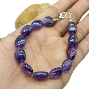 May include: A bracelet composed of smooth, oval-shaped purple amethyst beads strung together. The piece features a silver-toned metal lobster claw clasp at one end, providing a secure closure for the polished gemstone strand.