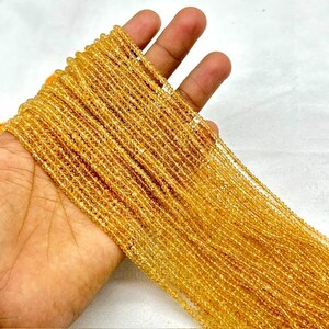 May include: A long strand of small, faceted, translucent yellow citrine gemstone beads. The beads are uniform in size and shape, strung together in a dense, continuous line, displaying a bright, golden-yellow hue throughout the entire length.