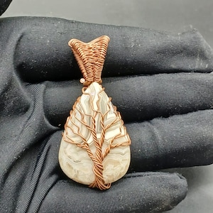 May include: A white agate pendant with a copper wire tree design. The pendant is shaped like a teardrop and has a copper wire bail.