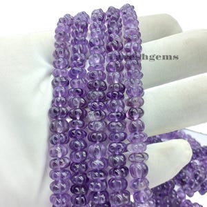 May include: Close-up of several strands of polished, translucent purple amethyst beads. Each bead features a carved floral design. The beads are strung together, suitable for a necklace or bracelet.