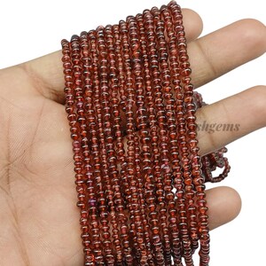 May include: Strands of small, round, dark red garnet beads. The beads are strung together, creating a flowing, textured appearance. The beads are held in a hand, showcasing their color and size. The word "shgems" is visible in the background.