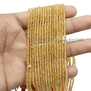 Puede incluir: Un hilo de pequeñas cuentas redondas de piedras preciosas de color amarillo dorado. Las cuentas están ensartadas, creando un efecto en cascada. Las cuentas se sostienen en una mano, mostrando su color y textura. El tema general es la fabricación de joyas.