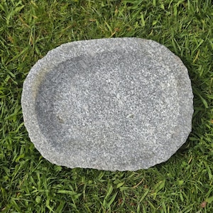 Può includere: Un piatto di pietra grigia di forma ovale con una superficie strutturata. Il piatto è posto su uno sfondo di erba verde. La pietra sembra essere granito o un materiale simile, con un motivo maculato.