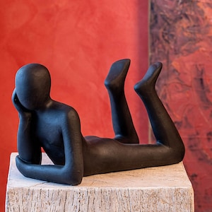 Può includere: Una scultura astratta nera di una persona sdraiata con la testa appoggiata sulla mano. La scultura è su una base di legno e su sfondo rosso.