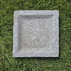Op de afbeelding: Een vierkante, grijze granieten stenen schaal met een iets verzonken midden. De steen heeft een gespikkeld patroon en ruwe randen. De schaal ligt op groen gras.