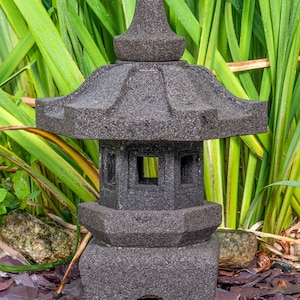 Könnte beinhalten: Eine graue Steinpagode-Laterne mit einem spitzen Dach und dekorativen Fenstern. Die Laterne ist von grünen Pflanzen und braunen Steinen umgeben.
