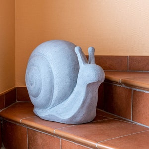 Può includere: Una scultura a forma di lumaca in cemento grigio chiaro con guscio e antenne dettagliate. La lumaca è posizionata su un gradino in piastrelle di terracotta, con un muro color pesca sullo sfondo. È un elemento decorativo da giardino.