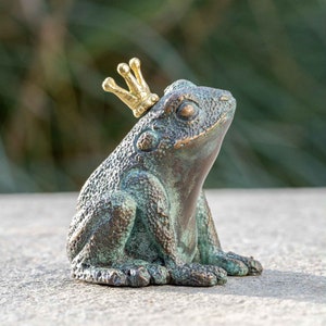 Può includere: Una piccola statuina di rana in bronzo che indossa una corona dorata. La rana è seduta eretta con le zampe ripiegate sotto di lei. La statuina ha una finitura patinata verde.