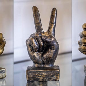 May include: Three gold-colored hand sculptures on wooden bases. The hands are making the OK sign, the peace sign, and the thumbs up sign.