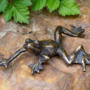 Può includere: Una scultura di rana in bronzo con una finitura testurizzata, in posa in posizione di salto su una roccia marrone. La rana ha un aspetto realistico con dettagli precisi.
