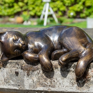 Op de afbeelding: Een bronzen beeld van een slapende kat, liggend op zijn zij met zijn poten onder zijn kin. Het beeld is gedetailleerd en realistisch, met een gladde, gepolijste afwerking.