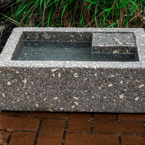May include: A rectangular, gray concrete birdbath with a small, rectangular ledge on one side. The birdbath is filled with water and is sitting on a brick patio.