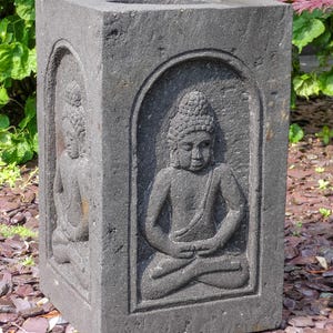 May include: A dark gray, rectangular stone planter with carved images of a seated Buddha in a meditative pose. The planter has a square opening at the top and is suitable for outdoor use. The Buddha figures are in a recessed arch design.