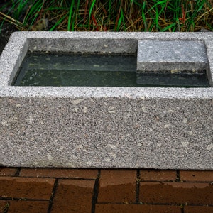 May include: A gray granite birdbath with a rectangular basin and a small ledge on one side. The basin is filled with water.