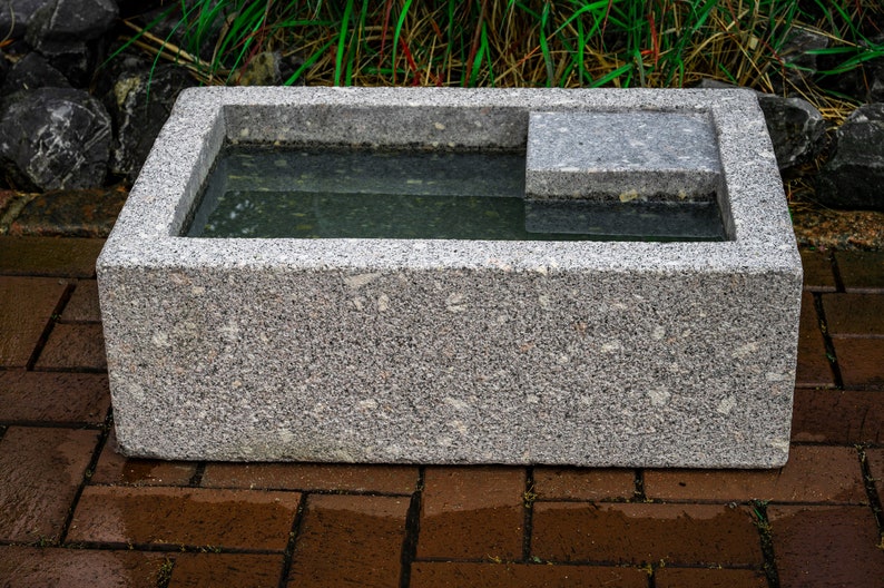 May include: A gray granite birdbath with a rectangular basin and a small ledge on one side. The basin is filled with water.
