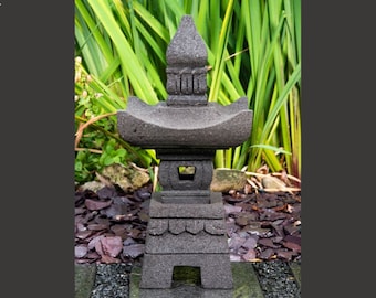 May include: A grey stone pagoda lantern with a tiered design. The lantern is sitting on a bed of dark grey gravel.