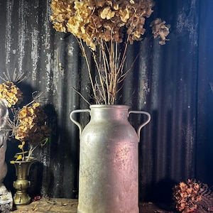 Puede incluir: Una antigua lechera de metal plateada con dos asas, llena de hortensias secas de color marrón claro. La lechera está sobre una superficie de madera rústica.