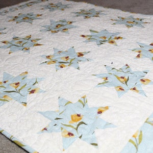 May include: A white quilt with a blue and yellow floral pattern. The quilt is made up of a series of star-shaped blocks.