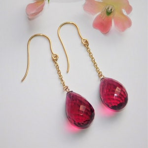 May include: A pair of gold dangle earrings with red teardrop-shaped gemstones. The earrings feature a delicate chain design.