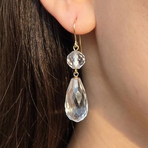 May include: A pair of gold-colored drop earrings. Each earring features a clear, faceted teardrop-shaped crystal pendant suspended below a smaller, round crystal. The earrings are hanging from a person's ear.