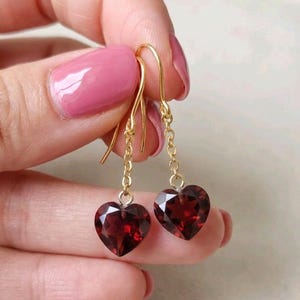 May include: Gold-colored dangle earrings featuring heart-shaped, faceted, deep red gemstones. The earrings have a delicate chain and hook closure. The gemstones are a rich, dark red color, and the metal has a polished finish.